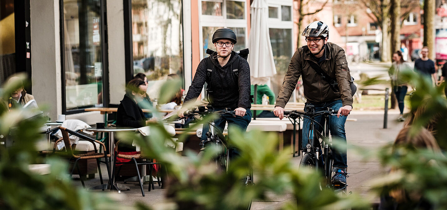 Zwei Männer mit Helm fahren auf einem Radweg im urbanen Raum